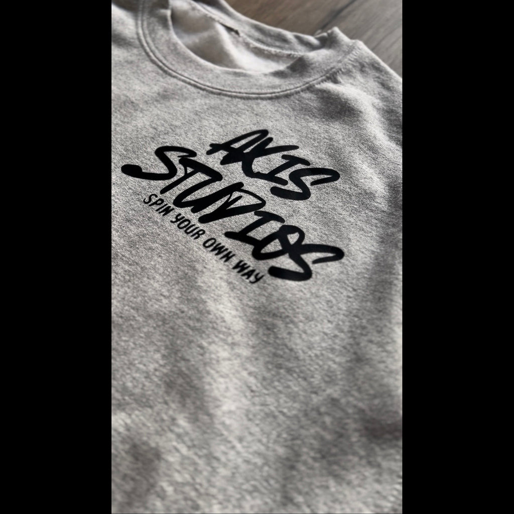 Close-up of a heather gray crewneck t-shirt featuring a black graphic print that reads 'AXIS STUDIOS' and 'SPIN YOUR OWN WAY!' in stylized lettering.