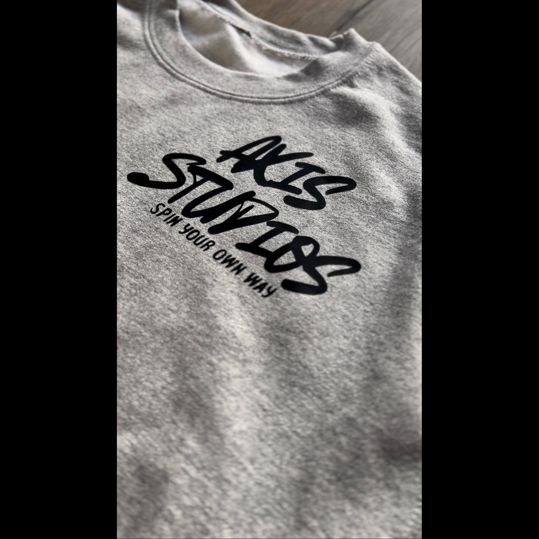 Close-up of a heather gray crewneck t-shirt featuring a black graphic print that reads 'AXIS STUDIOS' and 'SPIN YOUR OWN WAY!' in stylized lettering.