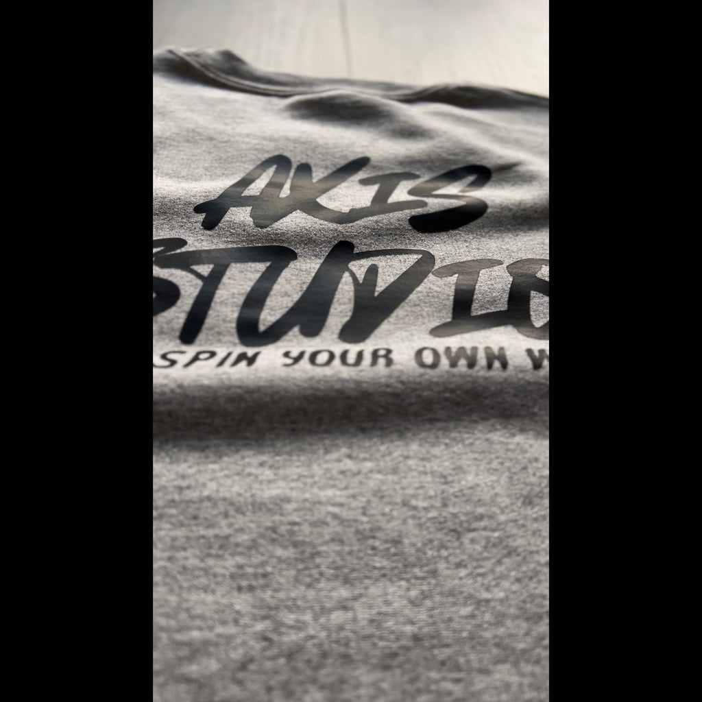 Close-up of a heather gray T-shirt with black printed text reading 'AXIS STUDIOS' and partial slogan text below, shown against a light neutral background.