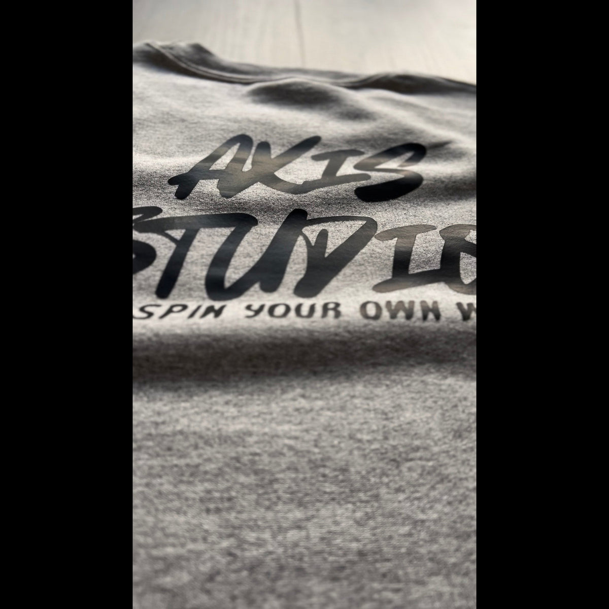 Close-up of a heather gray T-shirt with black printed text reading 'AXIS STUDIOS' and partial slogan text below, shown against a light neutral background.