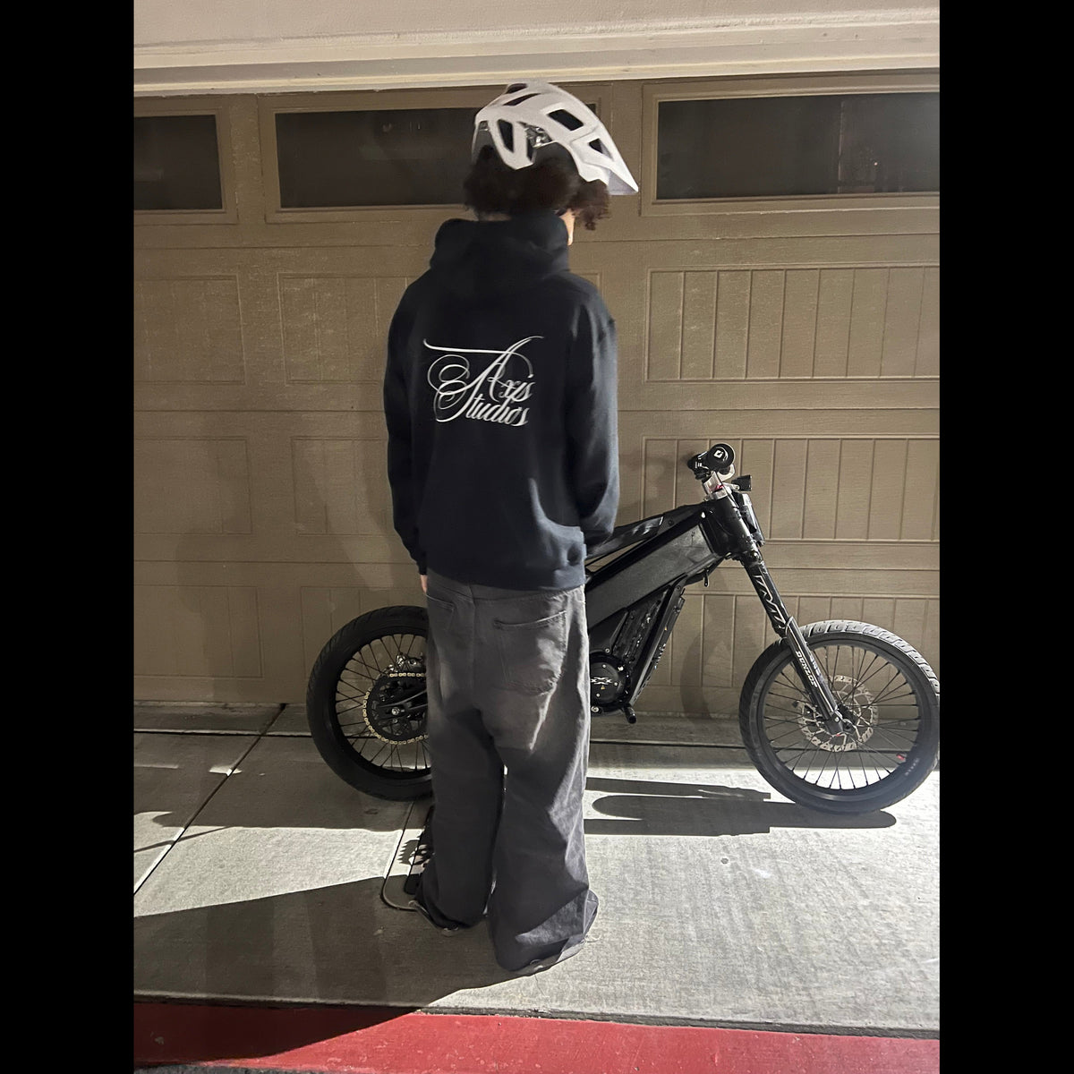 A person wearing a white helmet and a navy blue hoodie with a white graphic on the back stands next to a black electric mountain bike in front of a garage door.
