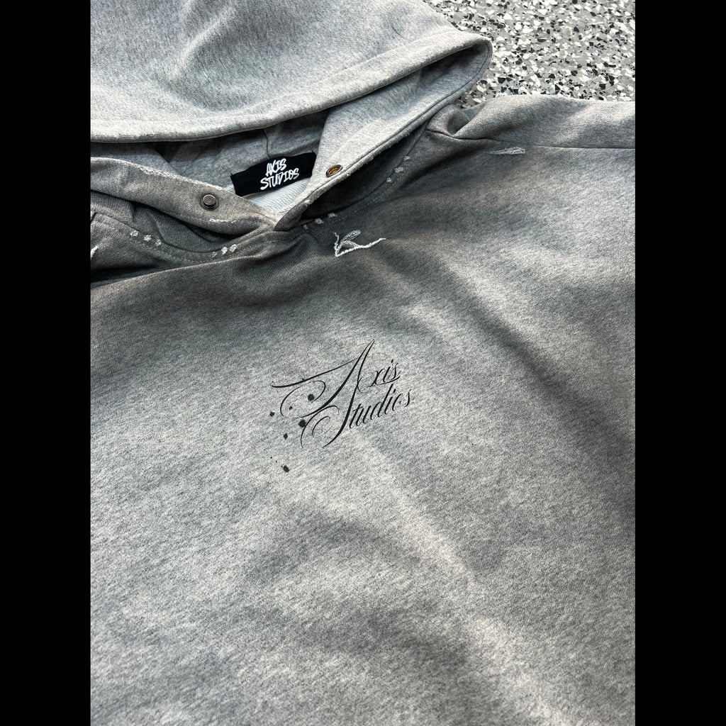 A close-up of a heather gray hoodie laid flat on a textured gray surface. The hoodie features a black embroidered script logo reading 'Art Studios' on the chest and a black brand tag inside the collar that reads 'Art Studios'.