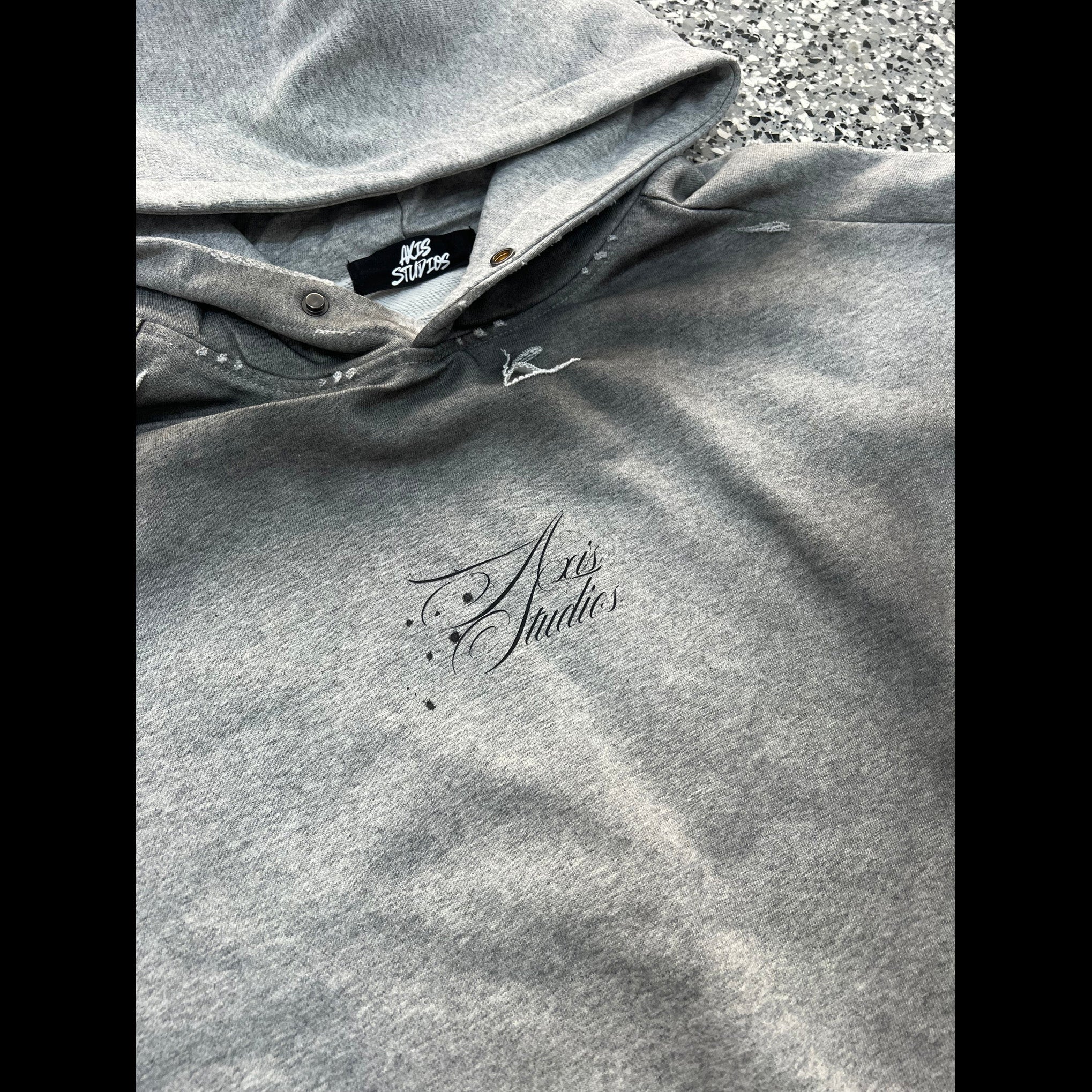 A close-up of a heather gray hoodie laid flat on a textured gray surface. The hoodie features a black embroidered script logo reading 'Art Studios' on the chest and a black brand tag inside the collar that reads 'Art Studios'.