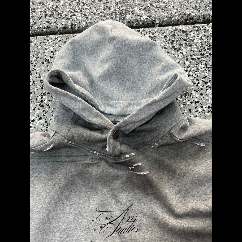 A close-up of a heather gray hooded sweatshirt laid flat on a speckled stone surface, with a small script logo near the lower front reading 'Axis Studios'. No person or real environment is present in the sense of a lifestyle scene; this is a product-focused flat lay, so subject_type is Product/Packaging.