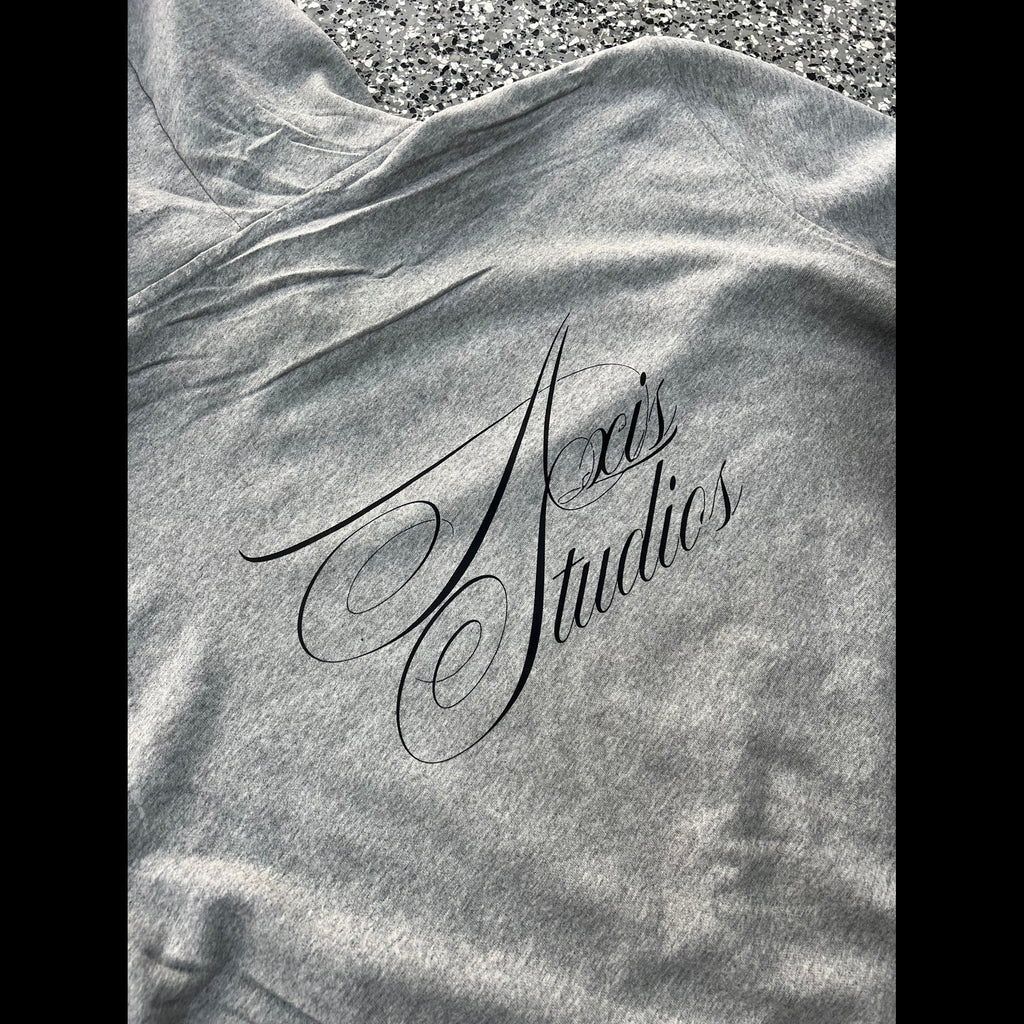 A close-up of a heather gray fabric item, likely a hoodie or sweatshirt, laid flat with the black script logo 'Axis Studios' printed across the chest. The fabric is wrinkled, and a gravel surface is visible at the top edge.