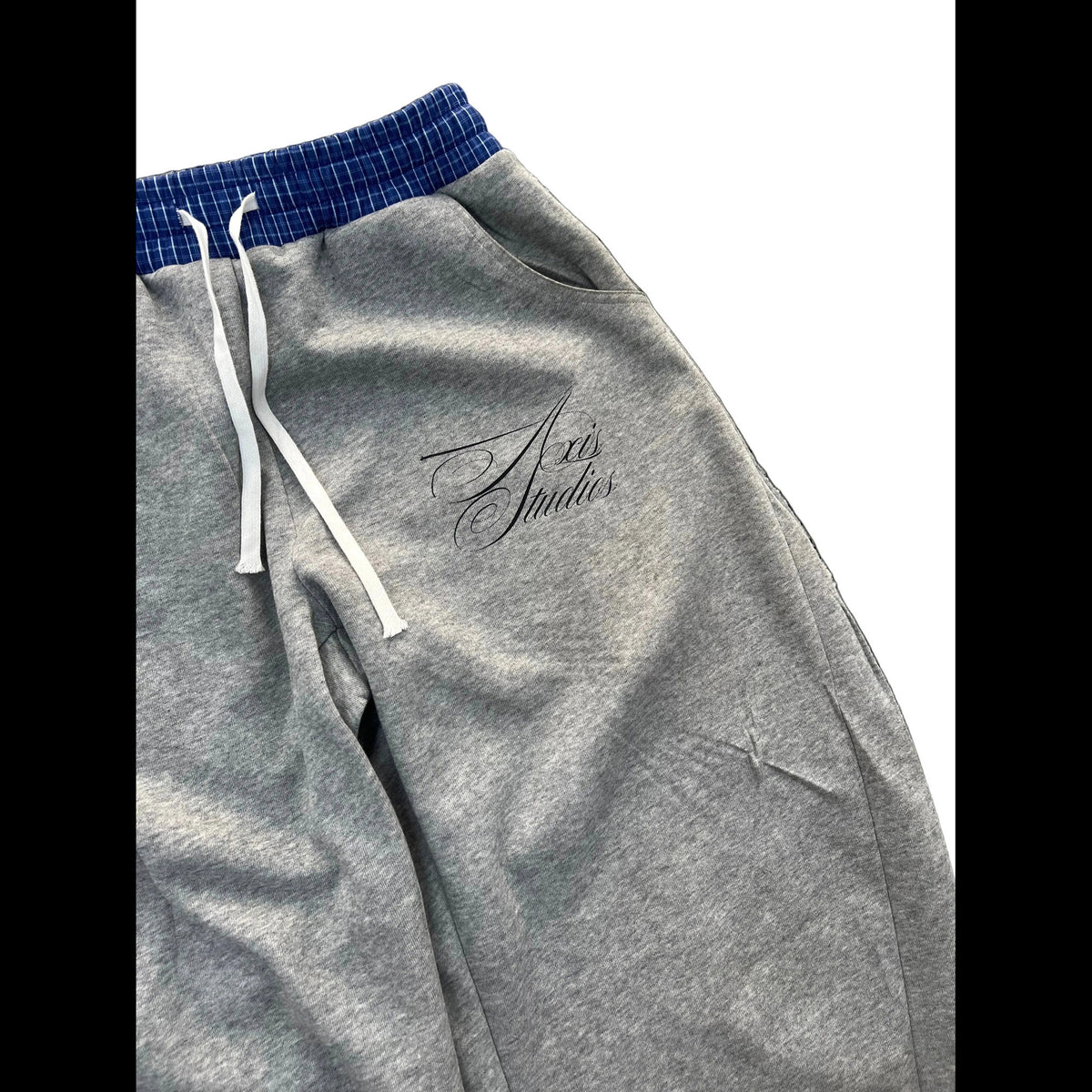 Close-up of heather gray sweatpants with a blue and white checkered elastic waistband, white drawstrings, and a black script logo reading 'A20 Studios' on the thigh.