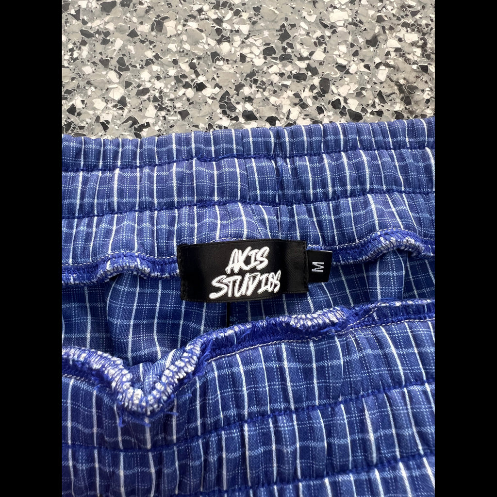 Close-up of blue and white plaid shorts showing the elastic waistband and a black fabric label that reads 'AC/BC STUDIOS' with a size 'M'.