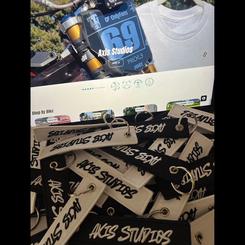 A pile of branded keychain fobs with metal keyrings, featuring black and off-white straps printed with 'AXIS STUDIOS' and 'SACRAMENTO BAY'. A motorcycle and part of a white T-shirt are visible in the background.