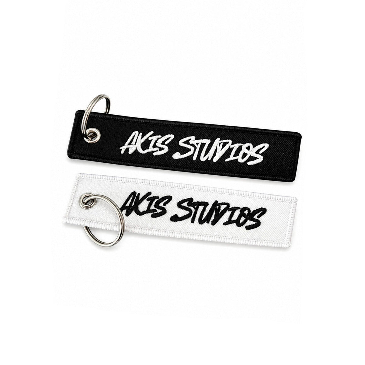 Two rectangular embroidered keychains or tags with silver split rings, shown on a white background. The top tag is black with white embroidered text reading 'AXIS STUDIOS', and the bottom tag is white with black embroidered text reading 'AXIS STUDIOS'.