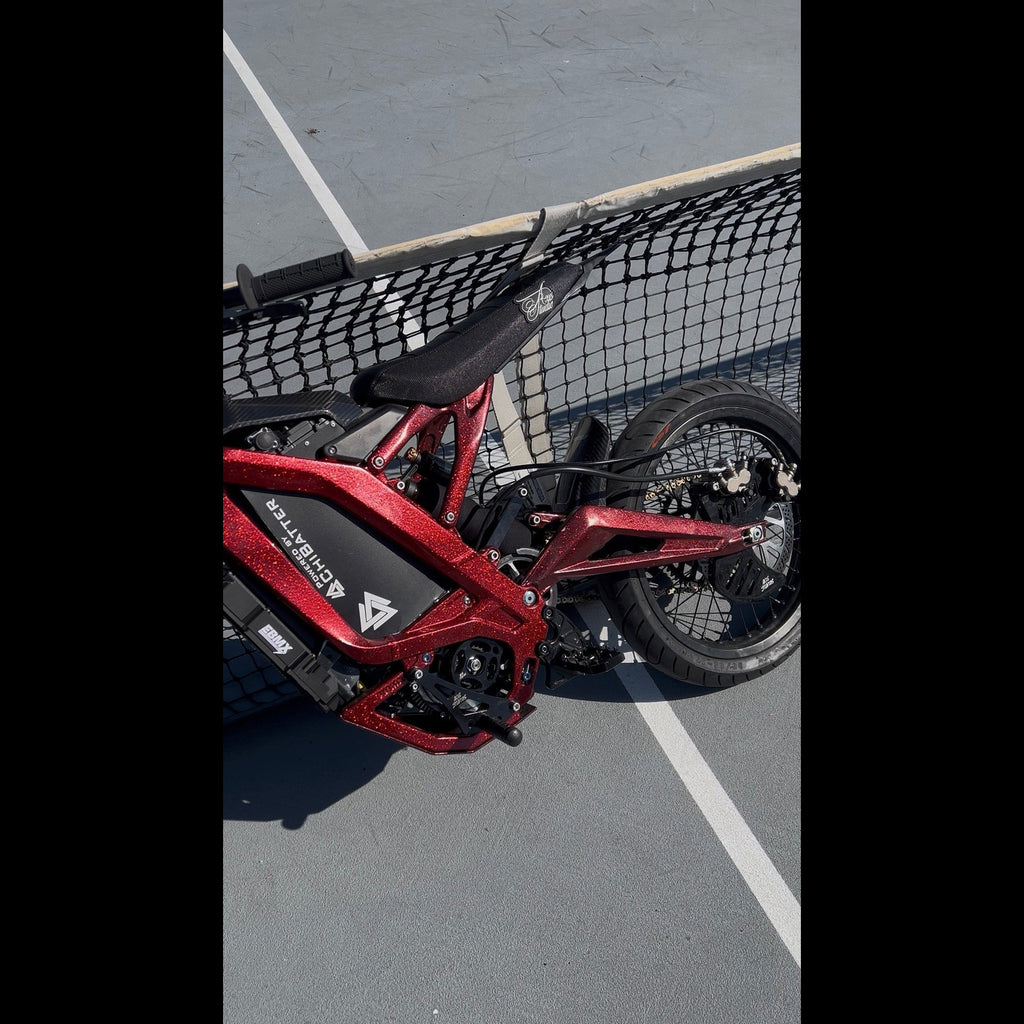 A red and black electric motorcycle with visible battery pack and rear wheel, photographed outdoors beside a black mesh fence on a gray paved surface.