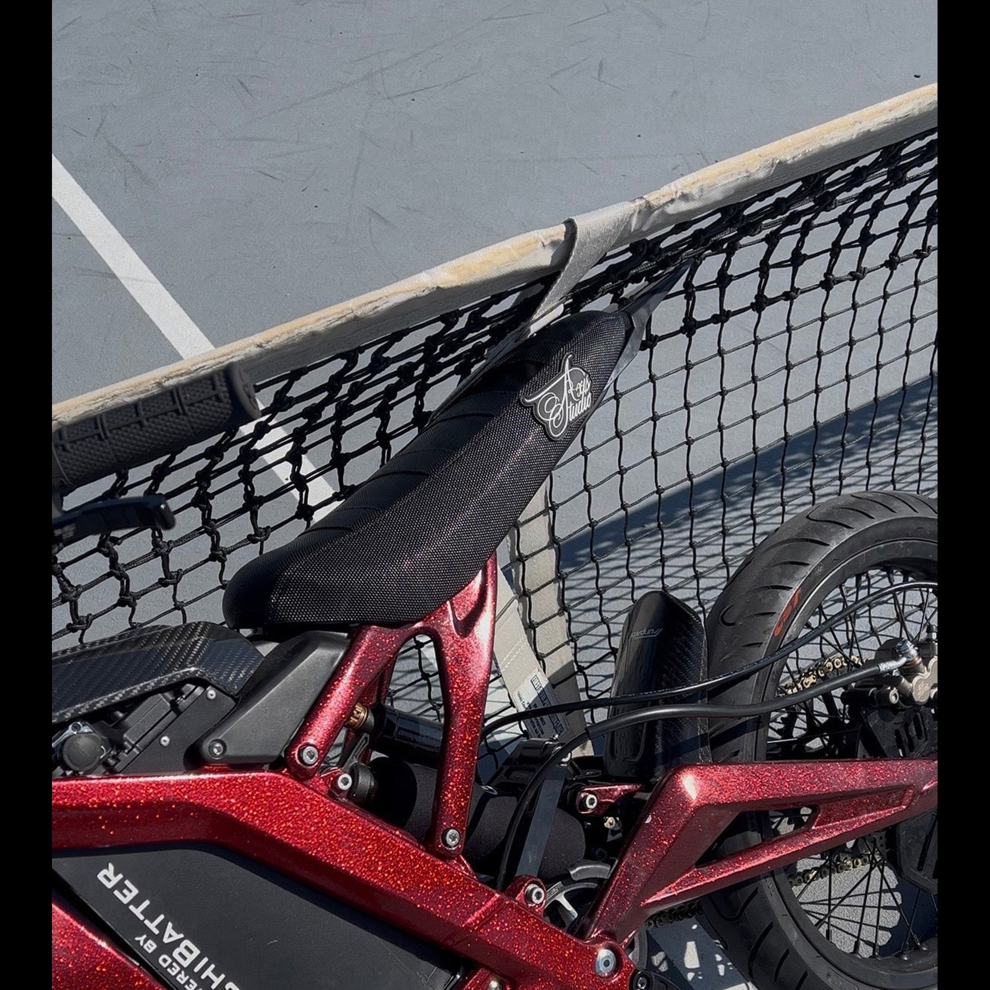 Close-up of a red electric bicycle frame with a black saddle featuring a white script logo, positioned behind a black mesh net on a gray paved surface.