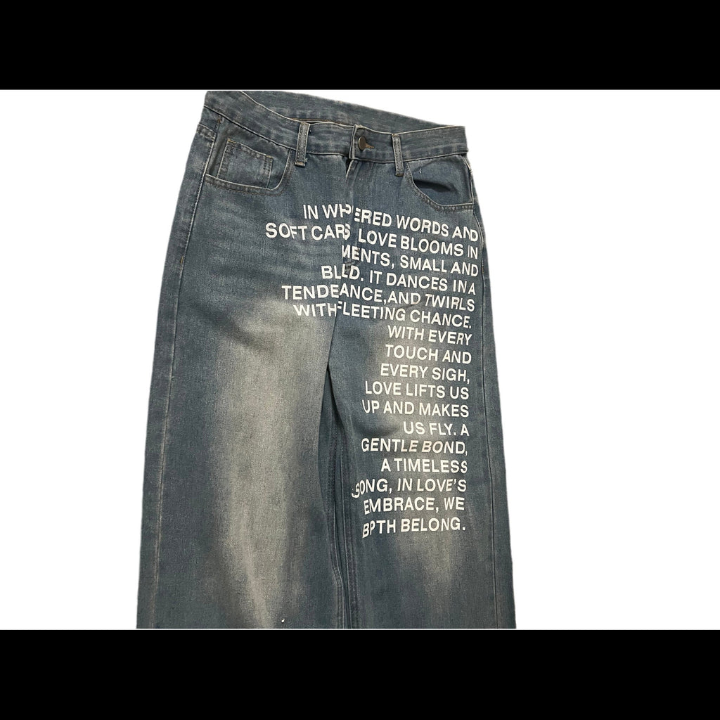A pair of faded blue denim jeans laid flat on a white background, rotated sideways. The front of the jeans features white printed text on the thigh.