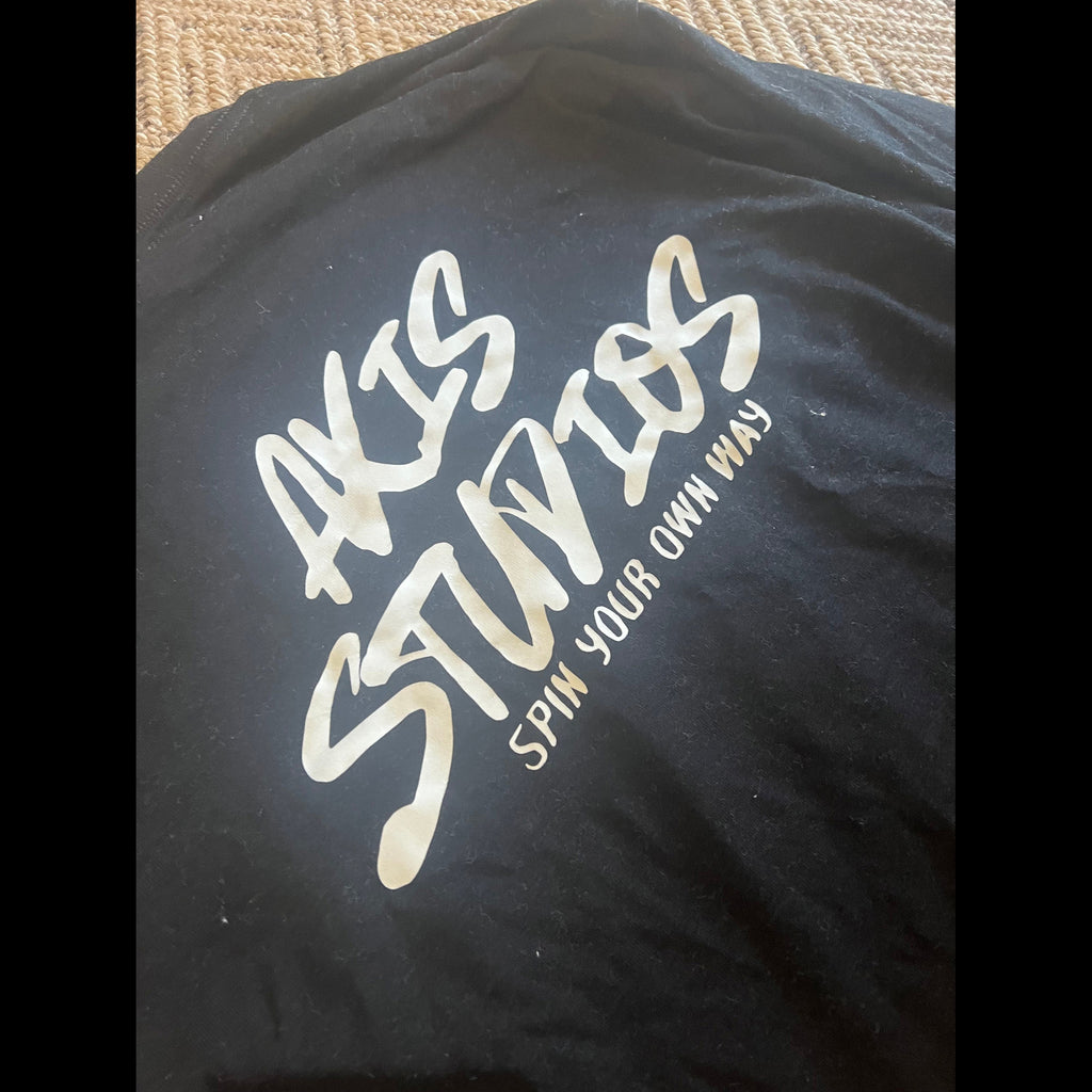 A close-up of a black T-shirt laid flat, featuring a large metallic gold foil print that reads 'AKIS STUDIOS' with the tagline 'SPIN YOUR OWN WAY' underneath.