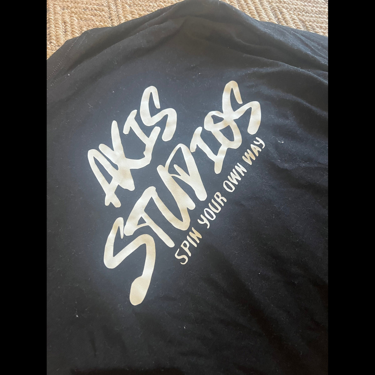 A close-up of a black T-shirt laid flat, featuring a large metallic gold foil print that reads 'AKIS STUDIOS' with the tagline 'SPIN YOUR OWN WAY' underneath.