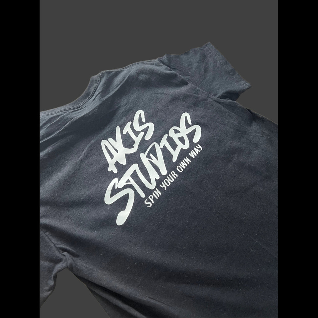 A close-up of a dark charcoal gray t-shirt featuring a white graphic print that reads 'AKIS STUDIOS' in a stylized script, with the tagline 'SPIN YOUR OWN WAY' underneath.