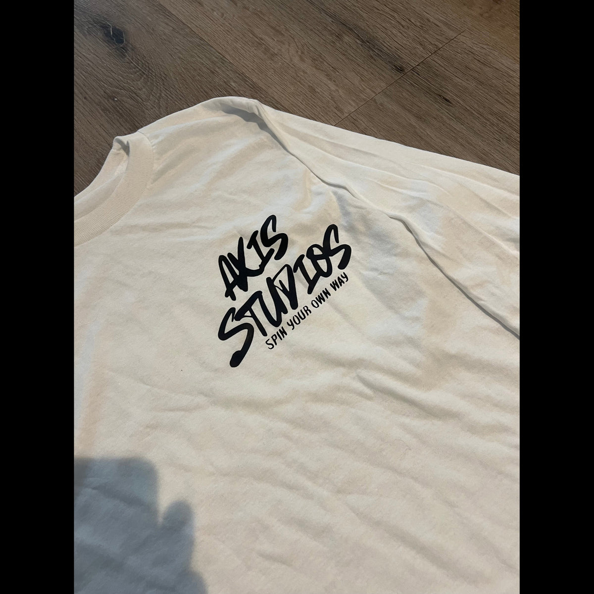 An off-white short-sleeve t-shirt with a black graphic print reading 'AKIS STUDIOS' and 'SPIN YOUR OWN WAY' on the chest, laid flat on a wooden floor.