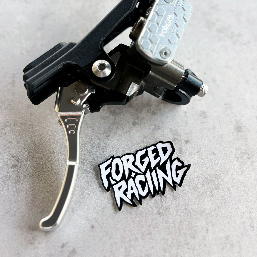 A close-up of a black and silver bicycle brake lever with a silver hex-patterned lever arm, placed on a light gray textured surface next to a black and white 'FORGED RACING' sticker.