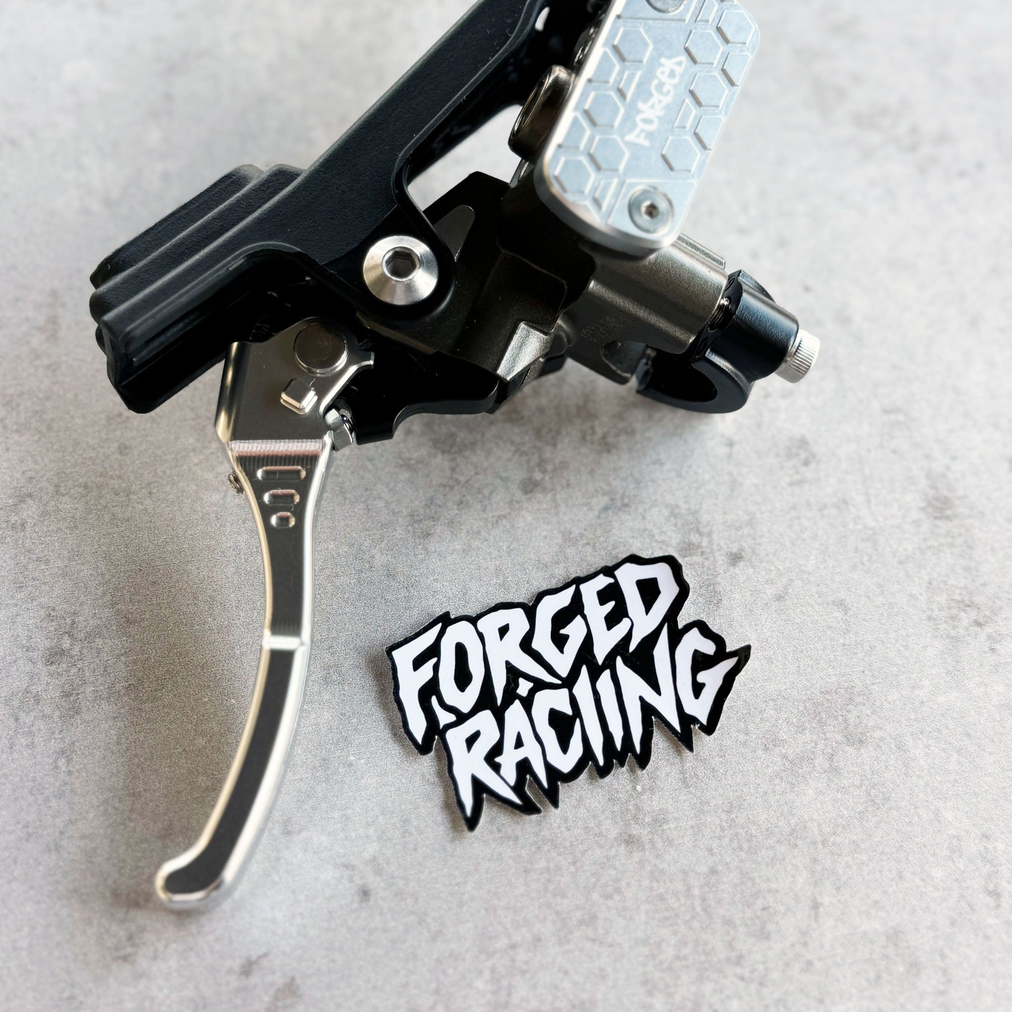 A close-up of a black and silver bicycle brake lever with a silver hex-patterned lever arm, placed on a light gray textured surface next to a black and white 'FORGED RACING' sticker.
