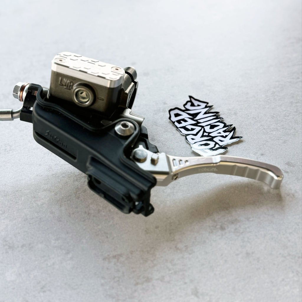 A close-up of a motorcycle brake lever assembly with a black lever body, silver metal brake cylinder and lever arm, shown on a light gray textured surface next to a black and white sticker.