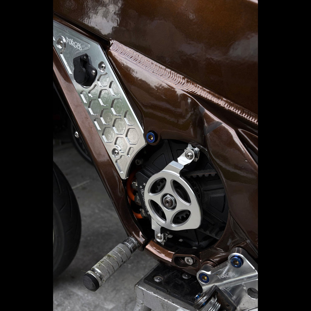 Close-up of a motorcycle's engine and side panel, showing a silver metallic chain guard with a honeycomb pattern and a silver sprocket wheel on a brown bike body.